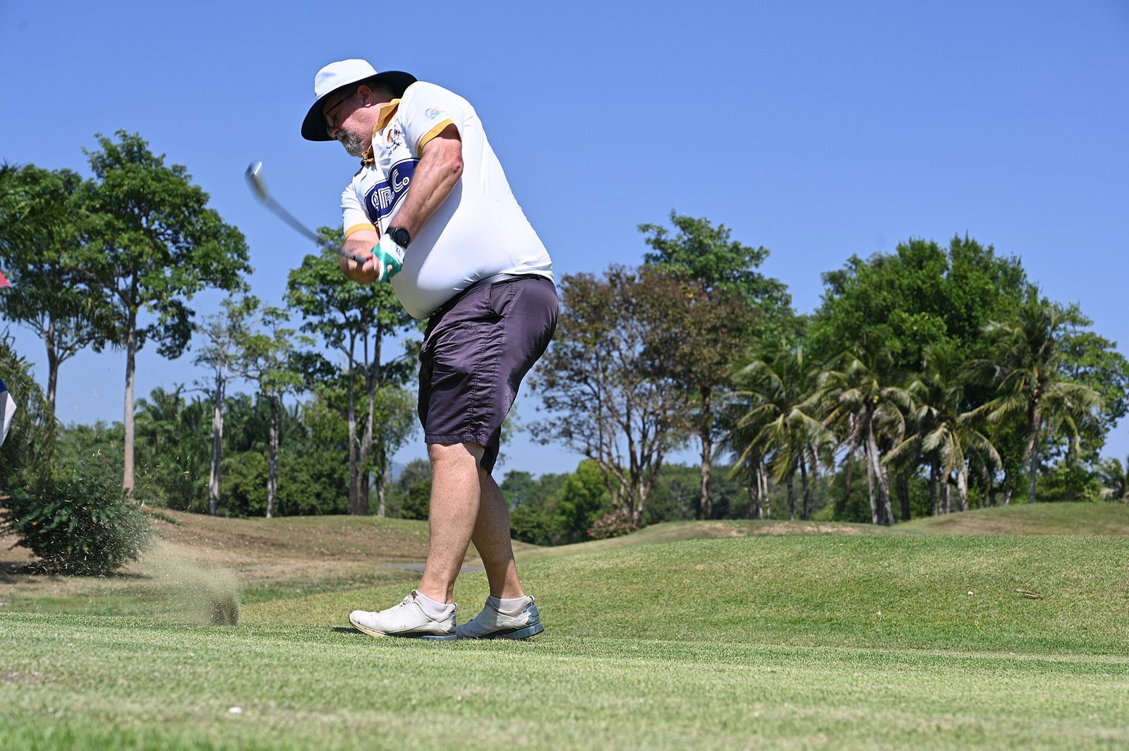 A golfer in a wide-brimmed white hat, white polo shirt and dark shorts swinging through a tee shot
